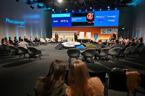gruppe von zuschauern auf der ITB Berlin im Zuschauerraum. In Front ein Vortragender neben einem Screenshot der Präsentation. Raum ist halbdunkel beleuchtet.